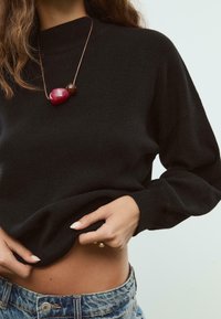 Žena u crnoj dukserici, plavim farmerkama i ogrlicom s dva velika perla, podiže duksericu da otkrije svoj struk.