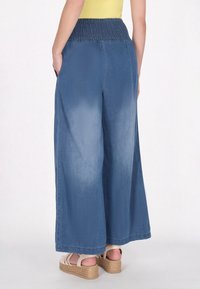 Sz éles szárú farmernadrág középkék színben, smockolt derékból, oldalsó zsebekkel és sima textúrával. A modellen bézs espadrille szandál van.