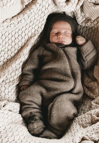 Rjavo otroško enodelno obleko iz flisa z zadrgo in pritrjeno kapuco, položeno na teksturirano kremno pleteno odejo.