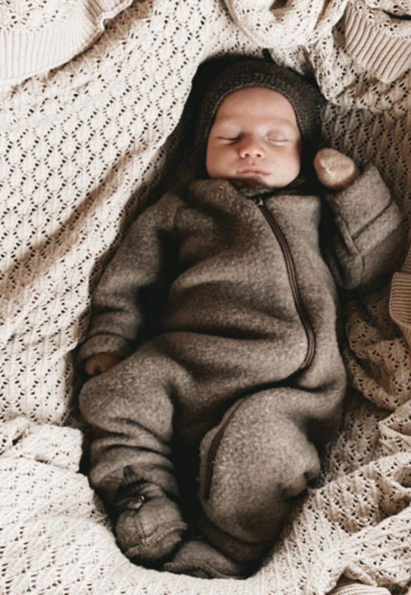 Rjavo otroško enodelno obleko iz flisa z zadrgo in pritrjeno kapuco, položeno na teksturirano kremno pleteno odejo.