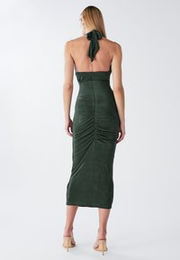 Zelena satenasta tesno prilegajoča obleka z zavezovanjem okrog vratu, dolžine do sredine meč, z naborki na hrbtni strani, kombinirana z bež čevlji na peto.