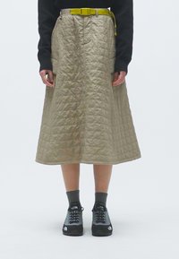 Saia bege acolchoada com um design de comprimento médio, apresentando um cinto amarelo brilhante. Ténis pretos e meias cinzentas completam o conjunto.