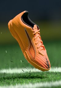 Orangener Sportschuh mit strukturiertem Obermaterial, kontrastierenden schwarzen Akzenten und detaillierter Schnürung, präsentiert über Gras mit verstreuten Rasenteilen.