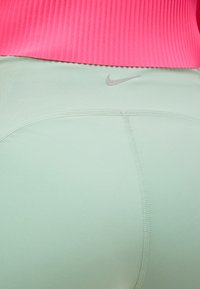 Κοντινό πλάνο σε ανοιχτοπράσινα κολάν Nike με ορατές λεπτομέρειες ραφών και έναν κοραλλί ριμπ αθλητικό τοπ πάνω από τη μέση.