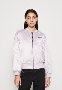 Giacca bomber di raso viola chiaro con zip frontale, polsini a costine e logo sul lato sinistro del petto; abbinata a jeans neri.