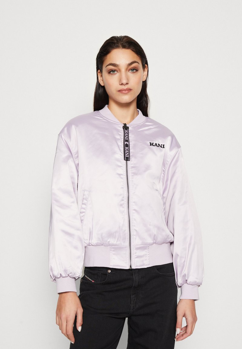 Giacca bomber di raso viola chiaro con zip frontale, polsini a costine e logo sul lato sinistro del petto; abbinata a jeans neri.