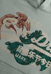 Design di felpa decorativa con un uccello, onde stilizzate e testo che recita "REALE" e "MARCA" abbellito con piccole borchie dorate.