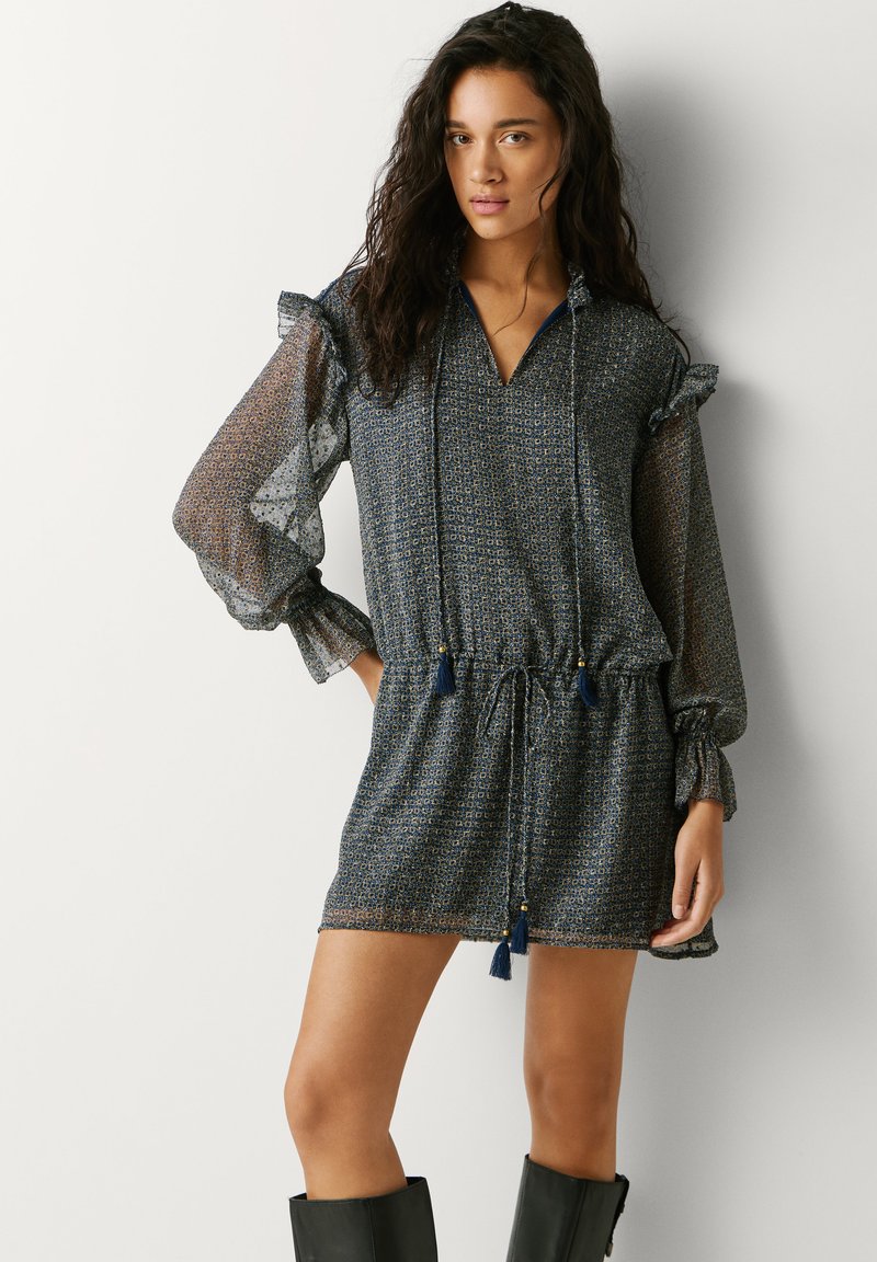 Vrouw draagt een blauw marine mini-jurkje met een patroon, doorschijnende pofmouwen en zwarte laarzen tot de knie, staand tegen een effen witte achtergrond.