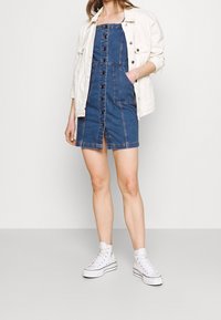 Kvinna klädd i en denimklänning med knappar, vit denimjacka och vita högskaftade sneakers, stående mot en enkel ljus bakgrund.