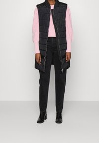 Gilet senza maniche imbottito nero sopra un maglione rosa chiaro testurizzato, abbinato a pantaloni neri e stivaletti neri lucidi.