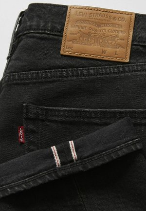 Jean en denim noir avec une patch en cuir marron présentant des détails en relief, un onglet rouge sur la poche et des accents rayés blancs et rouges au bas.