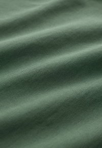 Material texturat de culoare verde, cu ondulații vizibile, realizat din bumbac moale, evidențiind o nuanță uniformă și un model de tricotaj fin.