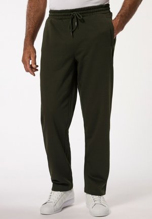 Mand iført mørkegrønne snøre bukser og hvide sneakers, stående med den ene hånd i lommen mod en ensfarvet baggrund.