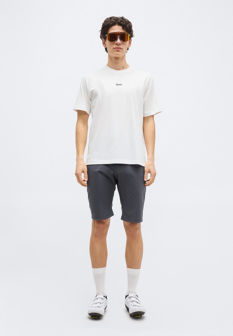 Man wearing white cycling shoes, white socks, gray cycling shorts, white Rapha t-shirt, and large orange-tinted sports sunglasses, standing.