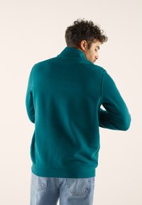 Suéter en color verde azulado, hecho de una tela suave, con cuello alzado, mangas largas y dobladillo acanalado. Usado por una persona de espaldas a la cámara.