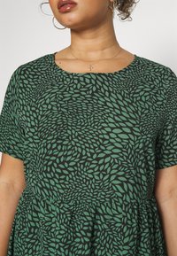 Vihreä lehtikuosi mustalla taustalla, lyhythihainen mekko pyöreällä pääntiellä ja kerätyllä vyötäröllä, valmistettu pehmeästä kankaasta.