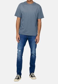 Blaues Baumwoll-T-Shirt mit Rundhalsausschnitt, kombiniert mit ausgefransten blauen Jeans und schwarzen Sneakers mit weißen Schnürsenkeln und Gummisohlen.
