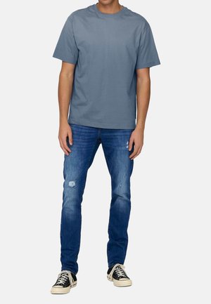 Blauw katoenen T-shirt met ronde hals, gecombineerd met versleten blauwe spijkerbroek en zwarte sneakers met witte veters en rubberen zolen.