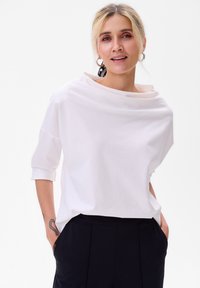 Witte, lichte, losvallende top met een gedrapeerde halslijn en driekwart mouwen. Gecombineerd met zwarte broek; minimalistisch ontwerp.