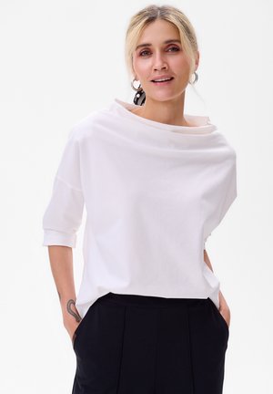 Haut blanc, léger, à coupe ample avec un décolleté drapé et des manches trois-quarts. Associé à un pantalon noir ; design minimaliste.