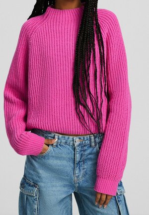 Personne portant un pull en grosse maille rose vif et un jean cargo taille haute bleu clair, une main dans la poche.