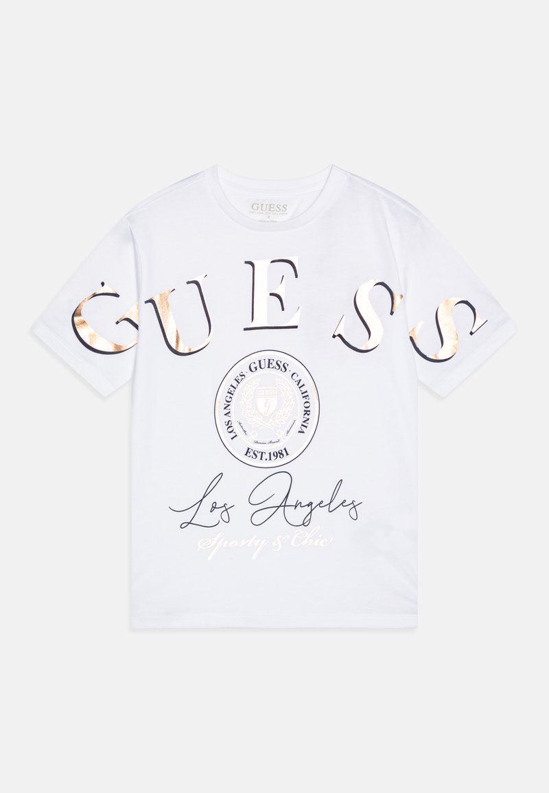 Bela bombažna majica s širokimi metalnimi črkami "GUESS", krožno logo s napisom "Los Angeles" in "Sporty & Chic" natisnjenim spodaj.
