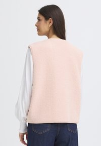 Rózsaszín gyapjú mellény, puha tapintású, kerek nyakkivágással és rögzítések nélkül, egy fehér hosszú ujjú ingre húzva, sötét farmernadrággal párosítva.