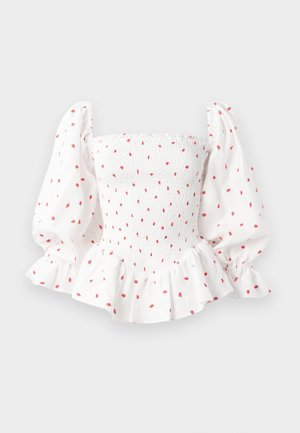 White blouse with smocked bodice, puffed sleeves, and ruffled hem. Patterned with small red dots. Cotton material with a soft texture.