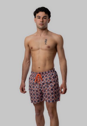 Giovane uomo in piedi senza maglietta, indossa pantaloncini da bagno con fantasia e coulisse arancione, su uno sfondo grigio uniforme.