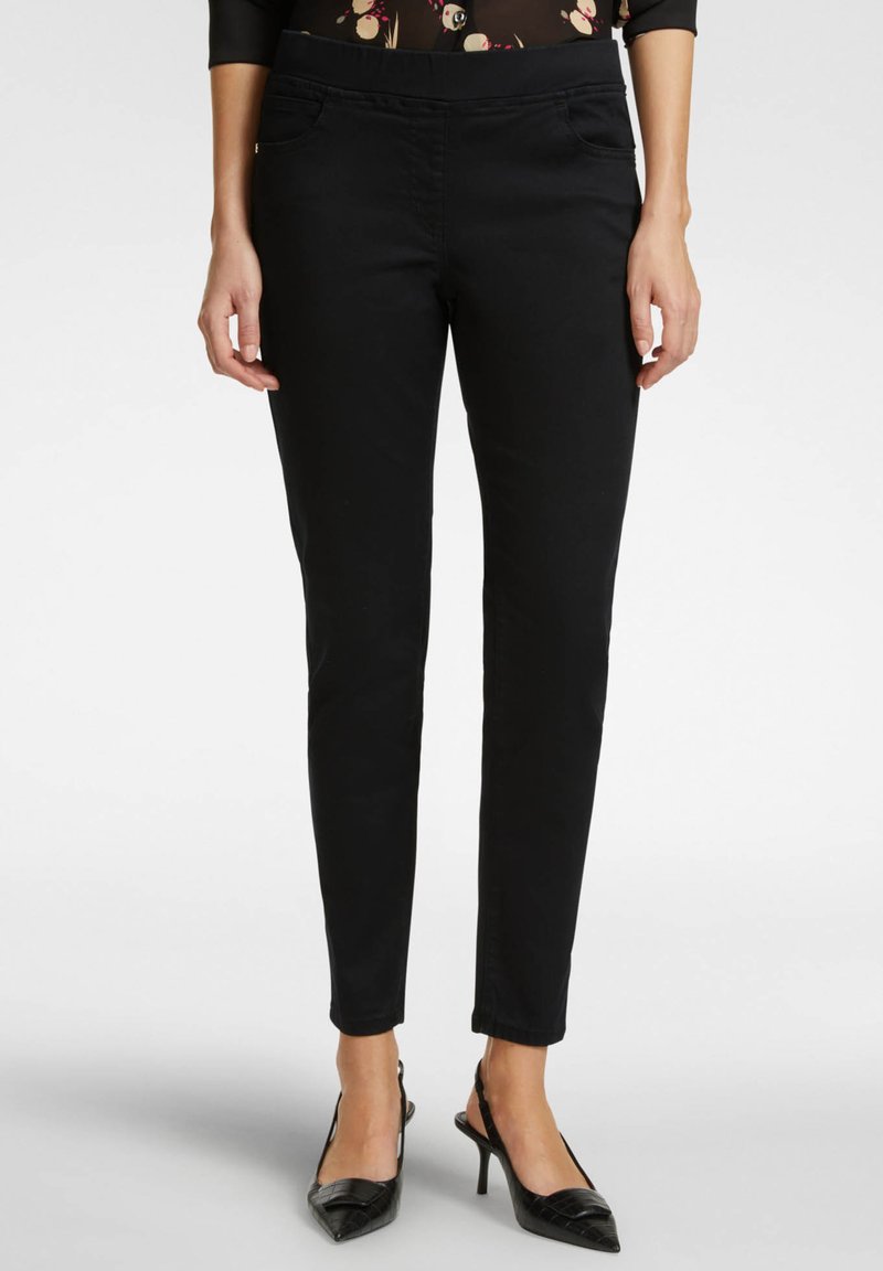 Pantaloni neri aderenti con una texture liscia, dotati di passanti per cintura e tasche frontali, abbinati a tacchi a punta neri.