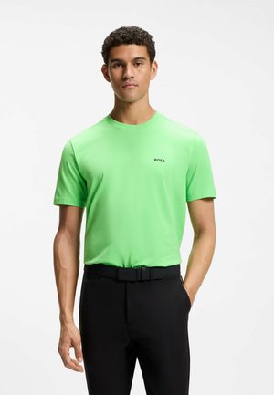 Junger Mann trägt ein hellgrünes BOSS T-Shirt, das in schwarze Hose mit schwarzem Gürtel gesteckt ist, und steht vor einem schlichten weißen Hintergrund.