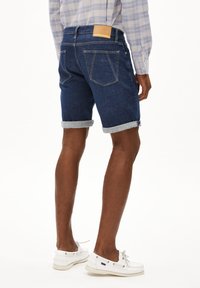 Donkerblauwe denim shorts met omgeslagen mouwen. Beschikt over twee achterzakken en een bruine leren patch. Gladde textuur, casual ontwerp.