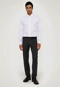 Bílá košile s límečkem na zapínání na knoflíky, doplněná tmavě šedými kalhotami. Oblečení zahrnuje černý pásek a lesklé tmavé boty.