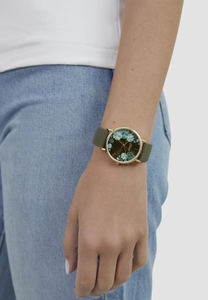 Runde Uhr mit goldenem Rahmen; grünem Silikonarmband; schwarzem Zifferblatt mit türkisfarbenem Blumendesign; schlankes Profil.