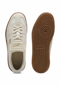 Białe skórzane sneakersy mają wyściełany język, teksturowaną powierzchnię oraz gumową podeszwę z okrągłymi wzorami. Widoczne jest logo "PUMA".