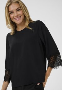 Schwarze Bluse mit Drei-Viertel-Ärmeln, die mit Spitzenbesatz an den Ärmeln, einem runden Ausschnitt und einer glatten Textur versehen ist.