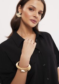 Czarna koszula z kołnierzykiem, złoty półksiężycowy bransoletka na nadgarstku i złote kolczyki w kształcie wachlarza. Widoczne gładkie tekstury i błyszczące wykończenia.