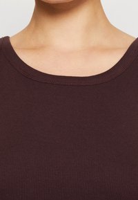 Burgunderfärgad ribbad linne med rund halsringning och strukturerad tyg. Synliga sömmar längs halsringningen ger extra detalj.