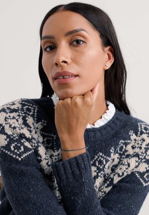 Tumszils adīts džemperis ar krēmkrāsas rakstainām detaļām un ribotām aprocēm, kopā ar baltu volānu apkakli. Models atbalsta zodu uz rokas.