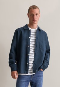 Navyblauwe blouse met knopen en een zak, gedragen over een witte gestreepte T-shirt, gecombineerd met lichtblauwe jeans. Soepele stof, casual pasvorm.