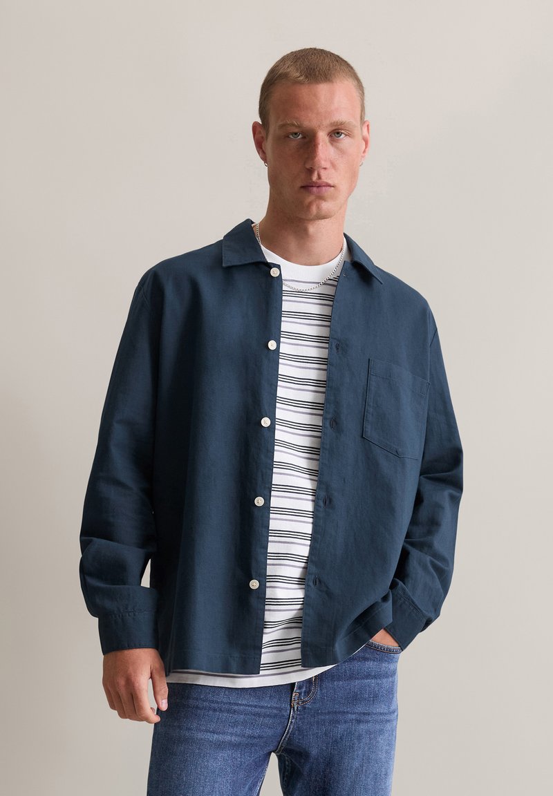 Navyblauwe blouse met knopen en een zak, gedragen over een witte gestreepte T-shirt, gecombineerd met lichtblauwe jeans. Soepele stof, casual pasvorm.