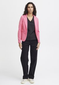 Blazer rosa in tessuto lavorato a maglia, abbinato a una maglietta nera con scollo a V e pantaloni neri a gamba larga. Sneakers grigie e bianche completano l'outfit.