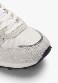 Zapatilla gris clara con parte superior de ante y malla texturizada, cordones blancos y suela de goma negra con detalles de costura sutiles.