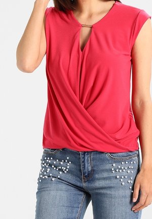 Kobieta nosząca czerwoną, bezrękawną bluzkę na zakładkę z metalową klamrą i niebieskie jeansy ozdobione małymi białymi perełkami na przednich kieszeniach.