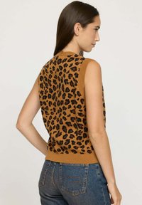 Femme de profil portant un haut sans manches à imprimé léopard brun et un jean bleu, sur un fond clair uni.