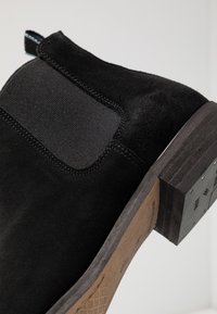 Bottine en daim noir avec panneau latéral élastique, talon carré, semelle texturée et tirant contrastant en teal à l'arrière.