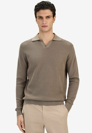 Mand iført taupefarvet strikket langærmet polo-sweater og beige bukser, stående med den ene hånd i lommen mod en hvid baggrund.