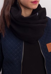 Black ribbed knit infinity scarf worn with a navy quilted jacket featuring a zip closure and a tan leather logo patch.