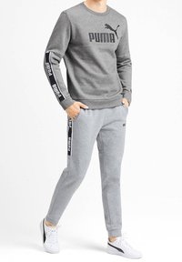 Šedá mikina a odpovídající tepláky vyrobené z měkké směsi bavlny; obsahují černé logo PUMA a branding podél rukávů a na boku kalhot. Bílé tenisky.