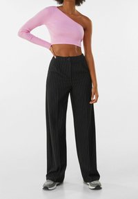 Top corto rosa acanalado de un solo hombro combinado con pantalones de pinstripe negros de pierna ancha. La modelo está de pie con una mano en la cadera, mostrando el atuendo.